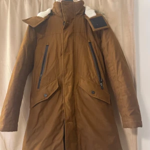 Brun parkasjacka med fuskpäls - Säljer en brun parkasjacka med huva och fuskpäls. Jackan har långa ärmar, dragkedjor på bröstfickorna och stora fickor med lock framtill. Perfekt för kalla dagar med sin varma och rejäla känsla. Snygg och praktisk design för vintern. Den är köpt för 8000 kr, och ännvänd fåtal gånger. Pris kan diskuteras! 💕