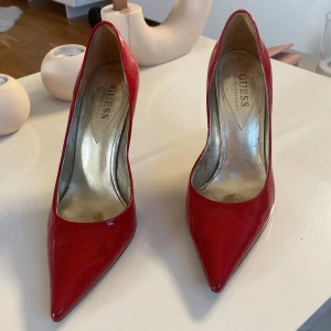 Röda pumps med spetsig tå från Guess - Snygga röda pumps från Guess med glansig finish och spetsig tå. Skorna har hög klack och en elegant siluett som verkligen sticker ut. Insidan är metallicfärgad och utsidan är i rött lackat material. Perfekta för dig som vill ha en statement-look.
