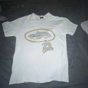 Vit t-shirt med guldkedje-tryck - Vit t-shirt i bomull med tryck av en guldkedja och motiv på bröstet. Klassisk passform och rund halsringning. Perfekt för dig som vill sticka ut med en unik och streetinspirerad stil. Har några svarta prickar som ej går bort i tvätten kan funka med bleknings medel men vet ej