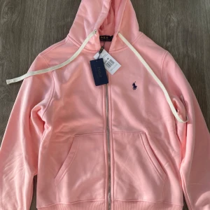 Rosa hoodie från Polo Ralph Lauren - Snygg ljusrosa hoodie från Polo Ralph Lauren med dragkedja framtill, huva med vita snören och klassisk mörkblå logga broderad på bröstet. Tillverkad i mjuk bomull och har känguruficka. Perfekt för en chill och stilren look.