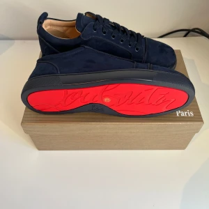 Mörkblå sneakers från Christian Louboutin - Snygga mörkblå sneakers från Christian Louboutin i mjuk mocka med klassisk röd sula och broderad logga på hälen. Skorna har rund tå, snörning och platt sula. Kommer med originalkartong, tre dustbags och äkthetskort. Priset kan sänkas vid snabb affär!