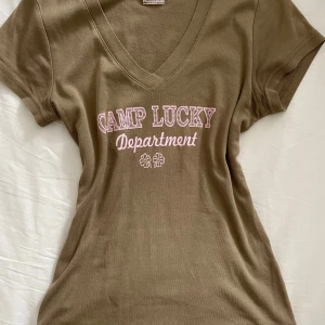 Vintage ONLY - Tröjan är från deras äldre kollektioner från 2000 talet. Svår att hitta. Klassisk V-ringning och korta ärmar. Den är figurnära vilket passar perfekt till low-rise jeans eller kjol Khaki/olivgrön färg med ljusrosa text.            Y2K, 2000-tal, Vintage, ONLY, Camp Lucky, Department, Olivgrön, Khaki, Rosa, Ljusrosa, Klöver, Slim Fit, Cropped, Magtröja, Ribbad, V-ringning, Retro, S (Small) #Y2K #y2kfashion #vintage #retro #ONLY #tshirt #topp #cropped #magtröja #khaki #olivgrön #rosa #pink #slimfit