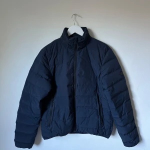 Mörkblå dunjacka från Henri Lloyd - Snygg mörkblå dunjacka från Henri Lloyd med hög krage och dragkedja framtill. Jackan har quiltad design och två fickor med dragkedja. Perfekt för kalla dagar och har en stilren look som funkar till det mesta. 3S6