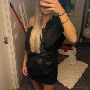 Svart glittrig one shoulder-klänning H&M - Svart kortklänning från H&M med glittrigt tyg och stor rosettdetalj framtill. One shoulder-modell💕endast använd vid ett tillfälle. Är 173 cm lång