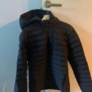 Svart dunjacka med huva från Peak Performance - Svart dunjacka från Peak Performance med huva och snygg quiltad design. Passformen är normal och jackan är långärmad.