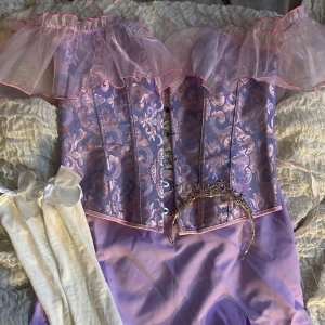 Lila prinsessklänning med volang - Superfin lila prinsesskorsett med glansigt mönstrad korsett, offshoulder volang i tyll och matchande kjol med slits. Inkluderar tiara med lila stenar och vita strumpor med rosetter. HELT OANVÄND! Allt i storlek M💖💖💓💓💓