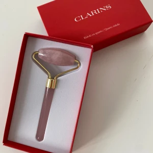 Rosa ansiktsroller i kvarts från Clarins - Säljer en lyxig ansiktsroller i rosakvarts från Clarins, aldrig testad. Rollern har ett elegant handtag i rosa kvarts och detaljer i guldmetall. Perfekt för att boosta hudvårdsrutinen och ge ansiktet en fräsch känsla. Kommer i snygg röd presentask.