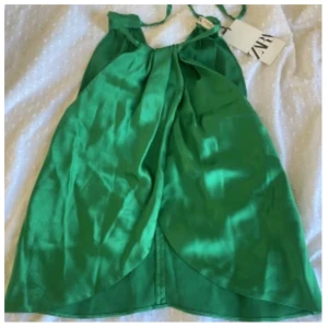 Grön omlott linnetopp från Zara - Snygg grön omlott-linnetopp från Zara med halterneck och knytning i nacken. Toppen har en glansig finish och är ärmlös, perfekt för varma dagar. Den har en unik omlott-design framtill som ger en cool och trendig look.