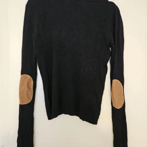 Svart kofta med mockadetaljer - Svart kofta från Laetitia Mem med v-ringning och knappar framtill. Långa ärmar med snygga bruna mockalappar på armbågarna. Mjuk och skön. Fint skick, knappt använd