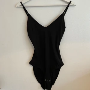 Svart body från MONKI med smala axelband - En stilren svart body med smala axelband och v-ringad urringning både fram och bak. Modellen är enkel och klassisk. Materialet känns mjukt och stretchigt, och underdelen har tryckknappar för enkel användning.