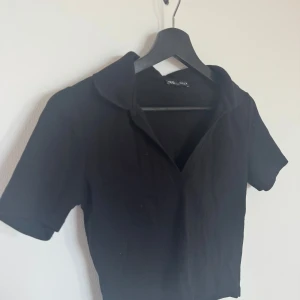Svart ribbad T-shirt med krage från Zara - Svart ribbad T-shirt med krage från Zara med korta ärmar och klassisk krage. Skjortan har en snygg v-ringning. Lite kortare i modellen 