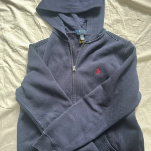 Mörkblå hoodie från Polo Ralph Lauren - Mörkblå hoodie från Polo Ralph Lauren med dragkedja och huva. Klassisk röd logga broderad på bröstet. Tröjan har långa ärmar och ribbade muddar. Perfekt för en chill och stilren look.