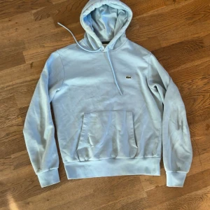 Ljusblå hoodie från Lacoste - Säljer en stilren ljusblå hoodie från Lacoste med klassisk krokodil-logga på bröstet. Tröjan har huva med snörning och en stor magficka framtill. Perfekt för dig som gillar en clean och avslappnad look.