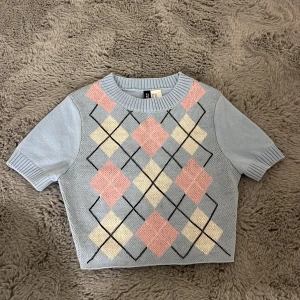 Ljusblå stickad tröja från H&M - Superfin ljusblå stickad tröja från H&M med korta ärmar och klassiskt argylemönster i rosa, vitt och svart framtill. Tröjan är croppad och har ribbade kanter vid hals, ärmslut och nederkant. Perfekt för en söt och trendig look!