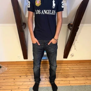 Mörkblå Polo Ralph Lauren Los Angeles t-shirt - Snygg mörkblå t-shirt från Polo Ralph Lauren med broderad Los Angeles-logga, stort vitt tryck och emblem på bröstet. Klassisk pikékrage, korta ärmar och nummerdetaljer på ärm och rygg. Slim fit-modell i bomull för en clean och sportig look. Påse följer även med. Storlek S.