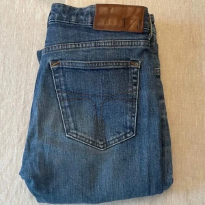 Blå jeans från T/J - Snygga blå jeans från T/J med klassisk femficksdesign och brunt läderpatch bak i midjan. Jeansen har raka ben och är tillverkade i slitstarkt denimtyg. Perfekta för en avslappnad och trendig look.
