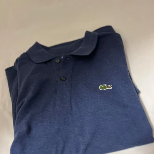 Blå pikétröja från Lacoste slim fit - Snygg marinblå pikétröja från Lacoste i slim fit-modell. Klassisk design med krage, två knappar och den ikoniska gröna krokodilloggan broderad på bröstet. Tillverkad i mjuk bomull som känns skön mot huden. Perfekt för en clean och sportig stil.