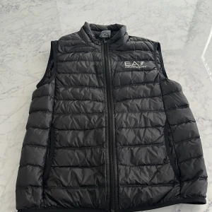 Svart dunväst från EA7 Emporio Armani - Snygg svart dunväst från EA7 Emporio Armani med quiltad design och dragkedja framtill. Västen har en hög krage och logga tryckt på bröstet. Perfekt för lager-på-lager och ger en sportig vibe.