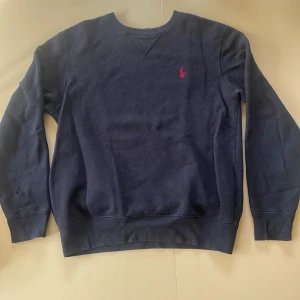 Svart tröja från Polo Ralph Lauren - Klassisk mörkblå tröja från Polo Ralph Lauren med rund hals och röd broderad logga på bröstet. Tröjan har långa ärmar och ribbade muddar. Perfekt för dig som gillar stilren och enkel design.