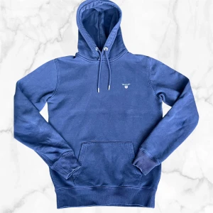 Blå hoodie från GANT med broderad logo - Snygg blå hoodie från GANT med broderad logo på bröstet. Tröjan har huva med snörning, känguruficka och ribbade muddar vid ärmslut och nederkant. Perfekt för chill dagar och enkel att matcha med jeans eller joggers. Jätte bra skick på tröjan. Några små slitmärken på ärmarna men syns inte(se bild) 