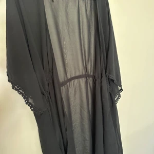 Svart transparent strandklänning - Svart strandklänning i tunn och transparent polyester med avslappnad passform. Klänningen har vida ärmar med dekorativ spetskant och små bollar längs kanterna. Perfekt för dig som gillar en bohemisk och chill vibe.