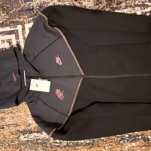 Svart och grå Nike hoodie med rosa detaljer - Nike hoodie i svart och mörkgrått med rosa loggor och sömmar. Tröjan har hel dragkedja, huva och två fickor med dragkedja. Perfekt för dig som gillar sportig och stilren streetwear. Storlek xl men känns som L