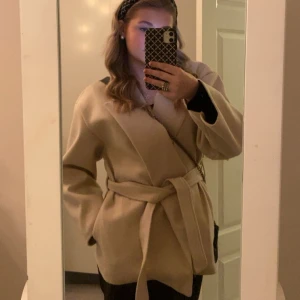 Beige omlottkappa med bälte - Säljer en beige kappa i omlottmodell med bälte i midjan. Jackan har en stilren och enkel design med långa ärmar och mjukt material. Perfekt för dig som gillar en clean och trendig look.