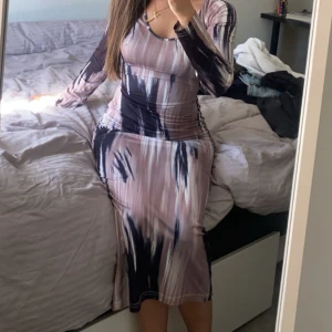 Långklänning med abstrakt mönster - Säljer en snygg långklänning med coolt abstrakt mönster i beige, svart och vitt. Klänningen har lång ärm, rund halsringning och ett knytband i midjan som ger en schysst siluett. Materialet är mjukt och stretchigt, perfekt för dig som gillar att sticka ut.