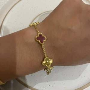 Guldarmband med röda fyrklöver - Superfint armband i guldton med fem fyrklöverformade länkar, varav tre är röda och två är helt i guld. Kedjan är tunn och smidig med justerbar längd och karbinlås. Perfekt accessoar för att lyfta din outfit med en färgklick och elegant design. Stål, guldpläterad och vattentålig.