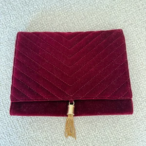 Vinröd sammet axelväska med guldkedja - Snygg vinröd axelväska i sammet med quiltad design och guldfärgad kedja. Väskan har ett dekorativt guldfärgat tofs-hänge framtill och stängs med lock. Perfekt för dig som vill sticka ut med en lyxig och trendig accessoar.