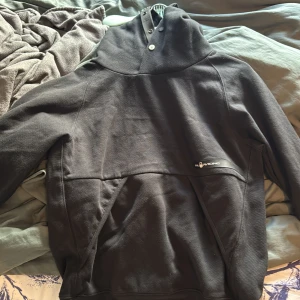 Svart hoodie från Sail Racing - Svart hoodie från Sail Racing med stor huva och snygga knappar vid halsen. Tröjan har en klassisk känguruficka framtill och är tillverkad i mjuk bomullsmix. Perfekt för dig som gillar stilren och sportig look.