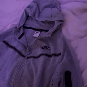 Grå Nike hoodie med dragkedja XL - Grå hoodie från Nike i storlek XL med svart Nike-logga på bröstet och svarta detaljer längs bröstet och på ena ärmen. Tröjan har huva, hel dragkedja och långa ärmar. Perfekt för chill eller träning.