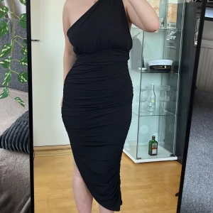 Svart oneshoulder klänning NA-KD - Säljer en svart, figurnära oneshoulder-klänning från Hanna Schönberg x NA-KD. Klänningen har asymmetrisk skärning, slits fram och snygg rynkning längs sidorna. Materialet är mjukt och stretchigt, perfekt för dig som vill sticka ut. Längd: 144cm. Byst: 40cm. 