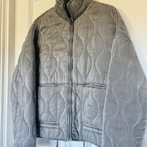 Grå quiltad jacka från Zara XS - Säljer en ny grå quiltad jacka från Zara i storlek XS. Jackan har quiltat mönster, dragkedja , hög krage och dragsko nertill. Materialet är mjukt. Finns en minimal fläck/märke, se bild 2