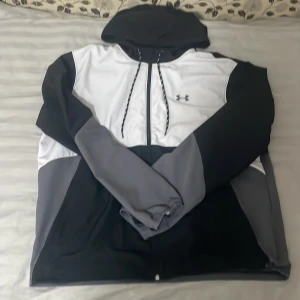 Under Armour hoodie i svart, grå och vit - Säljer en snygg hoodie från Under Armour i färgerna svart, grå och vit. Tröjan har huva med snörning, dragkedja framtill och långa ärmar. Materialet är polyester och insidan har meshfoder för extra komfort. Perfekt för dig som gillar sportig stil.