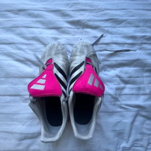 Adidas fotbollsskor vit/svart/rosa - Säljer ett par Adidas fotbollsskor i vitt med svarta ränder och detaljer i stark rosa. Skorna har snörning, klassisk design med täckt plös och dobbar i rosa undertill. Perfekta för dig som vill sticka ut på planen med både stil och funktion. Storlek 42