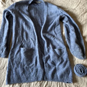 Blå stickad kofta med bälte - Mysig blå stickad kofta med långa ärmar och praktiska fickor framtill. Koftan kommer med ett matchande bälte för en justerbar passform. Perfekt för kyliga dagar!