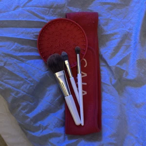 Sminkborstar och rengöringsplatta - Set med tre sminkborstar med vita handtag och silverfärgade detaljer, perfekt för applicering av puder, ögonskugga och blending. Medföljer gör en röd rengöringsplatta i silikon och ett rött fodral med Cala-logga. Allt du behöver för en flawless makeup-rutin.