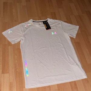 Under Armour T-shirt  - Snygg vit t-shirt från Under Armour med reflekterande logga. Modellen är kortärmad och har logga på bröstet samt text på ärm och nederkant. Tillverkad i lätt och ventilerande polyester. Hör av dig vid fler frågor eller funderingar.