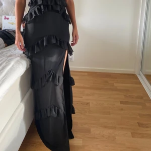 Svart maxi klänning - Säljer en svart axelbandslös långklänning från PrettyLittleThing, med flera lager volanger och en hög slits på ena sidan. Klänningen har en snygg twistad detalj vid bysten och sitter tight över kroppen. Perfekt till fest/bal och bröllop!!