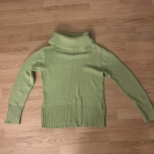 Ljusgrön stickad polotröja - Snygg ljusgrön stickad polotröja med bred ribbad krage och muddar. Tröjan har lång ärm och är perfekt för lager-på-lager under kyliga dagar. Enkel och stilren design som passar till många outfits. Små fläckar 