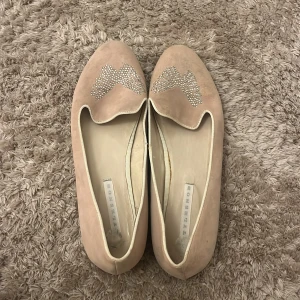 Ljus rosa ballerinaskor med strassdetalj - Snygga ljus rosa ballerinaskor i mocka med rund tå och glittrande strassdetaljer framtill. Skorna har en klassisk och stilren design som passar perfekt till jeans eller klänning. Mjukt material och platt sula för skön komfort hela dagen.