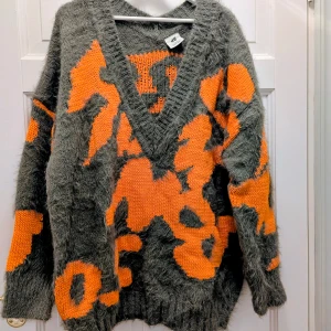 Grå-orange stickad tröja H&M Studio - Unik stickad tröja från H&M Studio AW2021 med coolt abstrakt mönster i grått och orange. Tröjan har en djup v-ringning, långärmade ärmar och en oversized passform. Materialet är fluffigt och mjukt, perfekt för att sticka ut med en färgstark vibe.