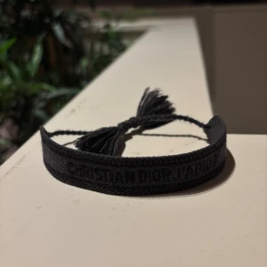 Christian Dior J'Adior armband - Ett svart tygarmband från Christian Dior med texten 'Christian Dior J'Adior' invävd. Armbandet har justerbar knytning med tofsar i ändarna och en stilren, minimalistisk look som passar perfekt till din outfit.