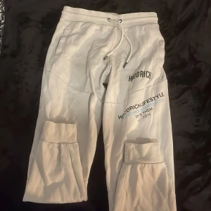 Vita joggers  från Hoodrich - Säljer ett par vita cargopants från Hoodrich med tryck på framsidan och stora fickor på benen. Byxorna har snörning i midjan och är tillverkade i mjuk bomullsmix för skön passform. Perfekta för en streetwear-look. Nypris 700