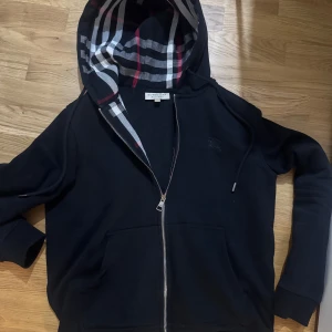 Burberry hoodie/kofta - Snygg svart hoodie fran Burberry med hel dragkedja och klassiskt rutigt foder i huvan. Broderad logga på bröstet och justerbar snörning i huvan. Perfekt för dig som vill ha en stilren och exklusiv look. Storlek M, passar även smal med en normal passform.