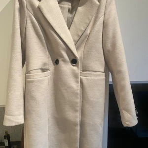 Beige kappa med knappar - Snygg beige kappa med klassisk krage och två svarta knappar framtill. Jackan har två fickor och är helfodrad. Långärmad och rak passform som ger en clean och stilren look. Perfekt för dig som gillar minimalistisk stil och neutrala färger.