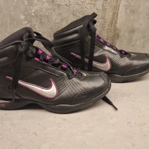 Svarta Nike sneakers med lila detaljer - Nike sneakers i svart med lila och silver detaljer. Modellen har högt skaft, snörning och en rund tå. Swoosh-logga . Skorna är tillverkade i syntetmaterial och har en sportig vibe. Kan användas både som träningsskor inne som ute.