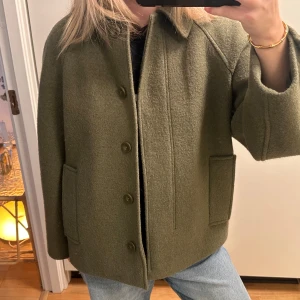 Olivgrön ulljacka med knappar - Grön kappa i ull från COS. Kappan har stora fickor framtill och stängs med knappar både fram och vid ärmslut. Som ny, använd fåtal gånger