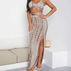 Beige omlott långklänning med slits - Snygg omlott långklänning i beige och bruna toner med randigt mönster. Klänningen har en öppen rygg, knyt i midjan och en hög slits på sidan. Toppen är ärmlös och har smala axelband som knyts i ryggen. Perfekt för dig som gillar trendiga och unika plagg.